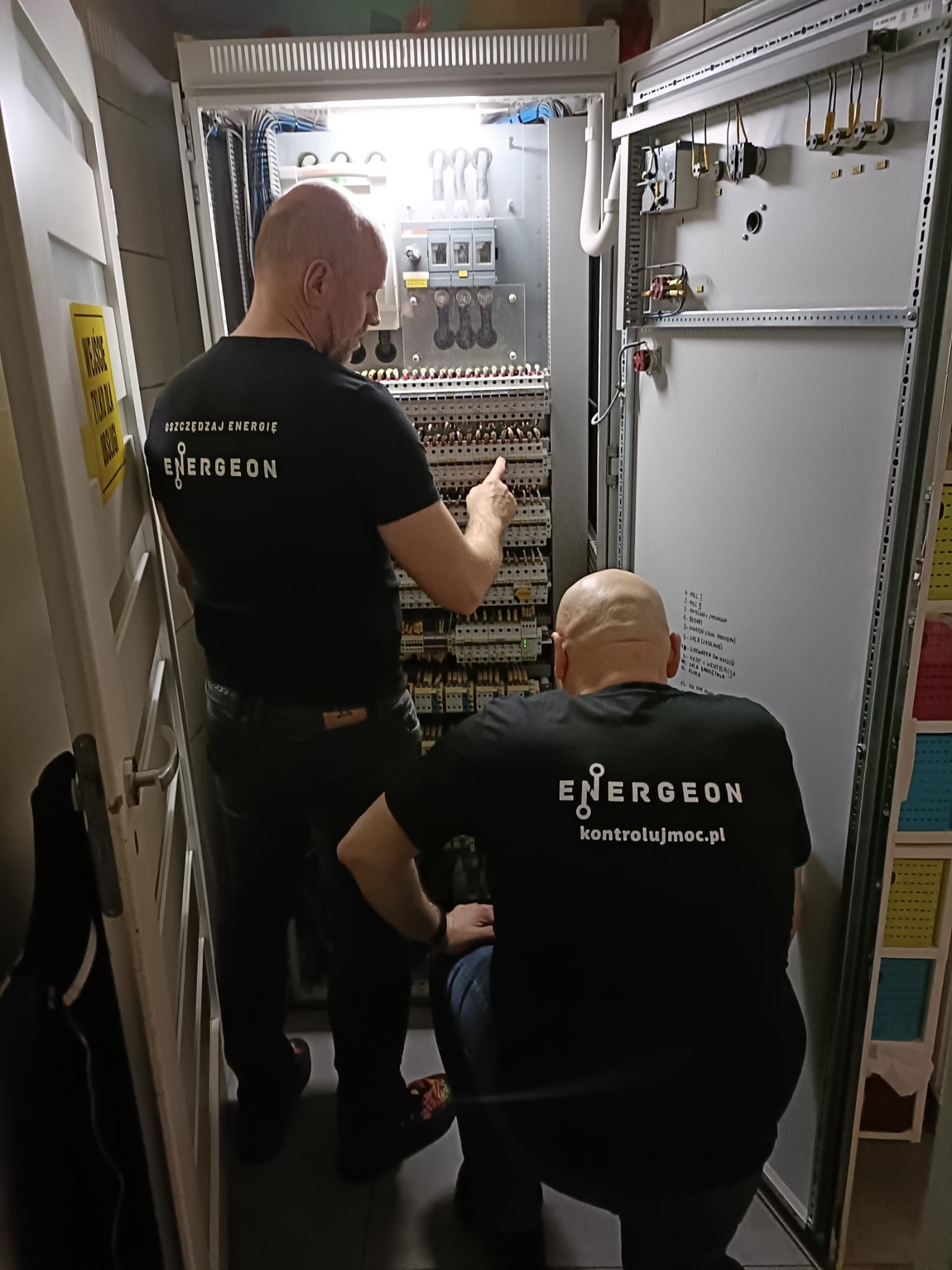 Dwóch techników ENERGEON w koszulkach kontrolujmoc.pl pracuje przy otwartej rozdzielnicy